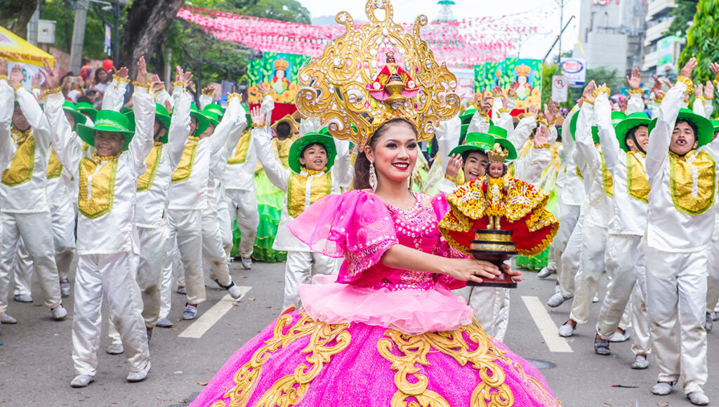 A vibrant image from the Sinulog festival showing a woman in a large pink gown and gold crown holding a Sto. Niño statue, surrounded by dancers in white and green costumes with raised hands.