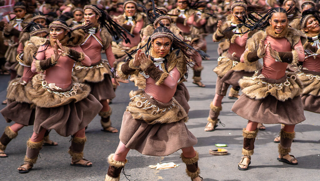 A dynamic scene from the Sinulog festival featuring a group of dancers in indigenous, fur-trimmed brown and pink costumes with braided hair performing a street routine.