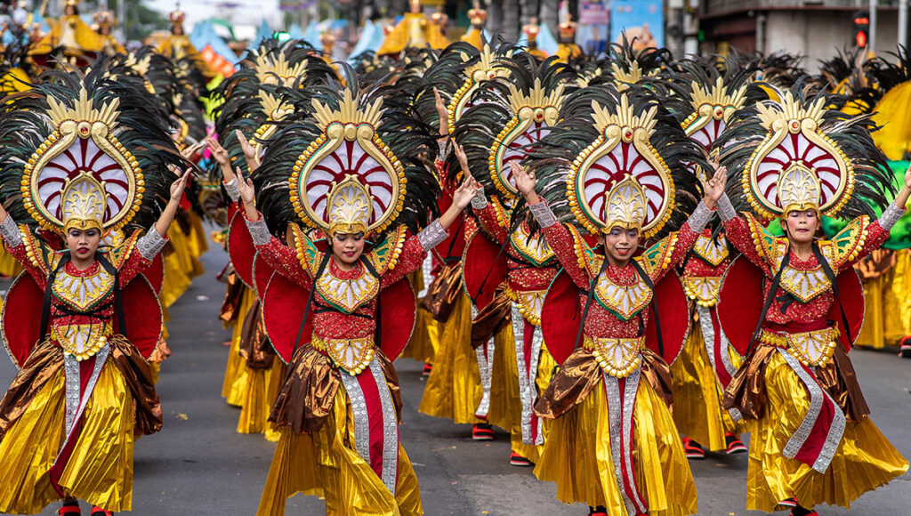 A massive line of performers at the Sinulog festival wearing elaborate red, gold, and brown costumes with large, black-feathered headdresses while dancing with arms raised.