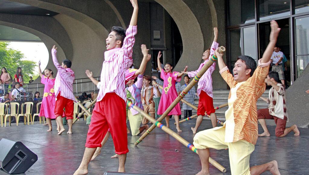 Filipino traditional dance called Tinikling performed by dancers in Manila, Philippines, showing performers skillfully stepping and hopping between clapping bamboo poles while wearing colorful traditional costumes during a cultural festival.