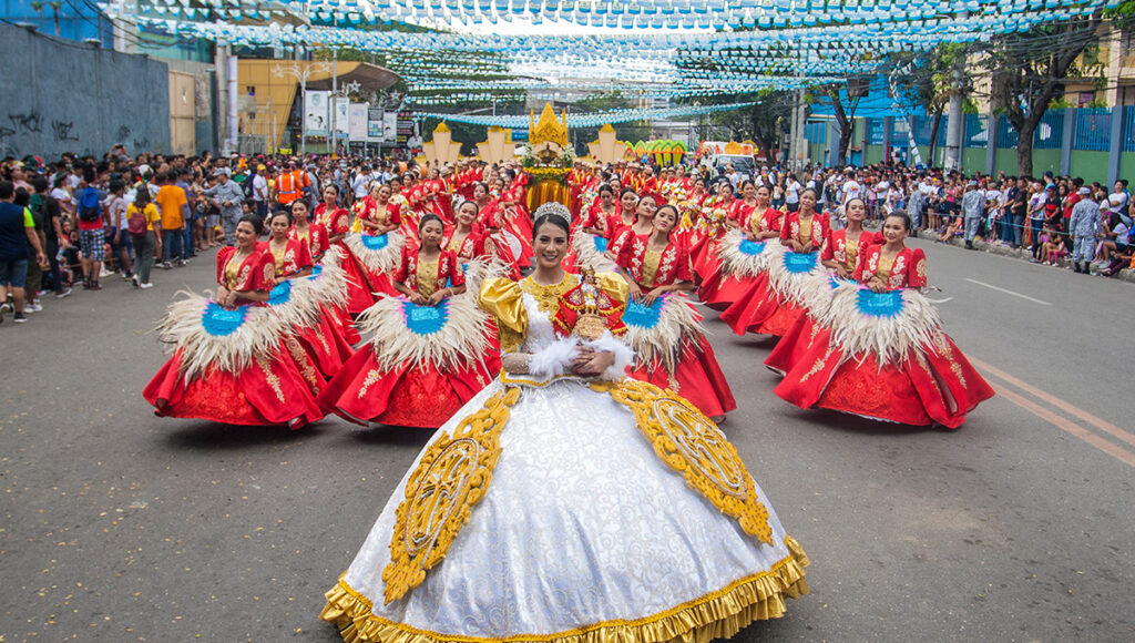 A central dancer in a large white and gold gown leads a group of female performers in voluminous red and blue costumes down the street during the Sinulog festival parade.