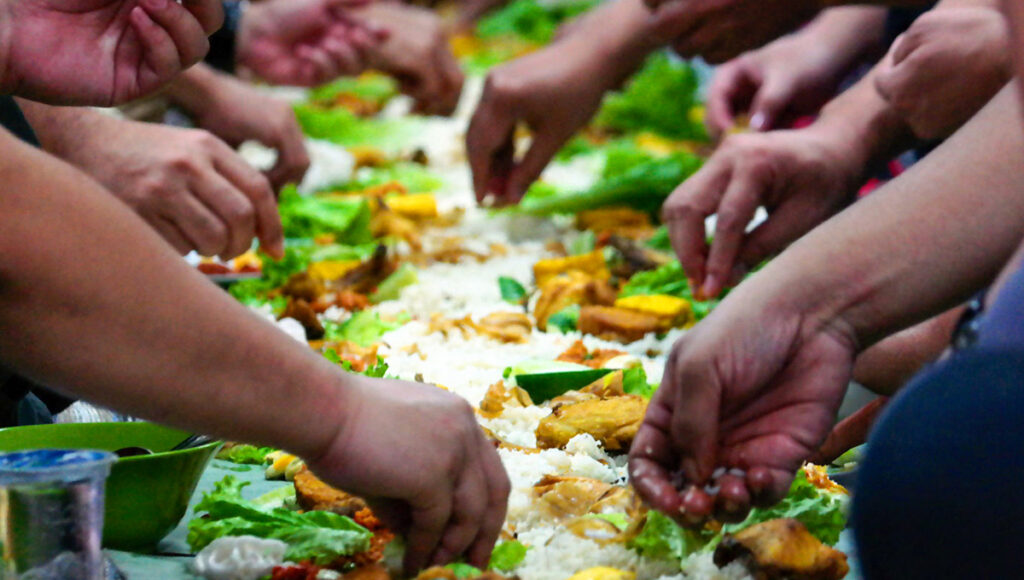 Guests enjoying a traditional Boodle Fight Philippines, eating with their hands from a communal spread of rice and assorted Filipino dishes on banana leaves.