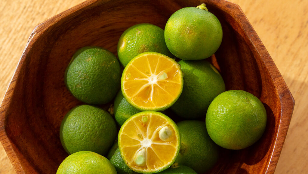 Top‑down view of a bowl filled with fresh green calamansi fruit halves arranged on a wooden surface.