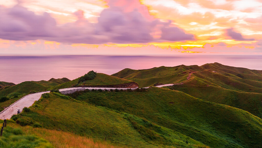 This breathtaking Batanes tourist attraction shows the famous Vayang Rolling Hills with smooth green slopes meeting sharp coastal cliffs. The landscape appears layered and wide, with lush grassy ridges leading the eyes toward the blue sea below, creating a striking contrast between land and ocean that makes Vayang one of the most scenic viewpoints in Batanes.