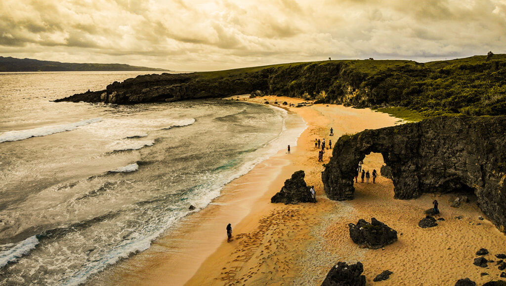 This must-see Batanes tourist attraction shows Morong Beach with the famous Nakabuang Arch, a large rock formation rising beautifully near the shoreline. The beach looks calm and inviting with fine white sand, gentle waves, and an open horizon, making it one of the best swimming spots in Batanes and a perfect place for relaxation and island photos.