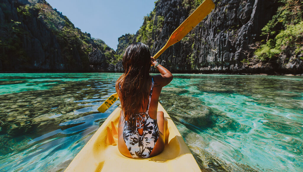 A woman kayaking through crystal clear tropical waters between limestone cliffs, a key activity to consider when deciding how long to spend in the Philippines