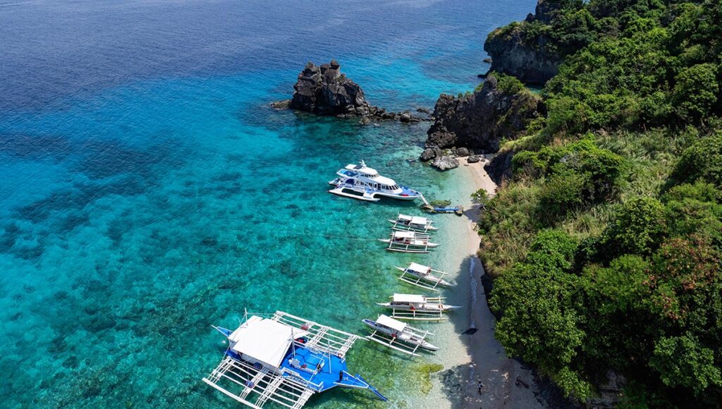 A high-angle view of white outrigger boats and a yacht anchored in clear blue water next to a hidden beach, showcasing island hopping tours offered by family resorts in Philippines.