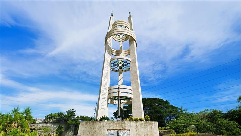 A striking view of the Philippine-Japanese Friendship Tower, a key landmark among historical places in Bataan. The 27-meter-tall white monument consists of three soaring pillars interconnected by several circular rings, symbolizing the renewed bond between Japan and the Philippines. Set against a bright blue sky, the tower stands at a major road junction in Bagac as a symbol of peace and reconciliation following World War II.
