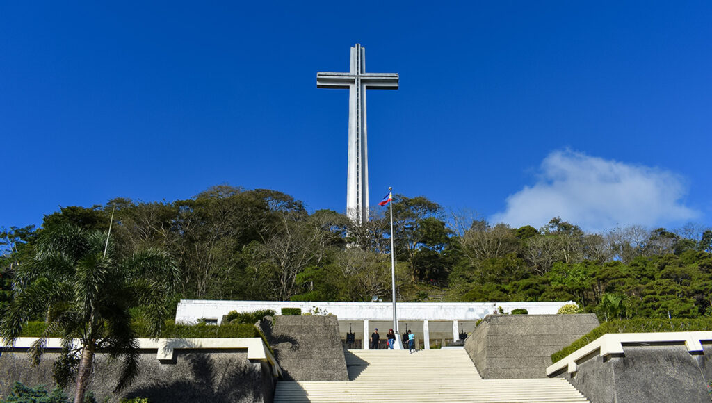 A towering white memorial cross at the Mount Samat National Shrine, one of the most prominent historical places in Bataan. The massive structure stands atop a lush, tree-covered hill under a clear blue sky, with wide stone steps in the foreground leading up to a white colonnade where visitors are walking.