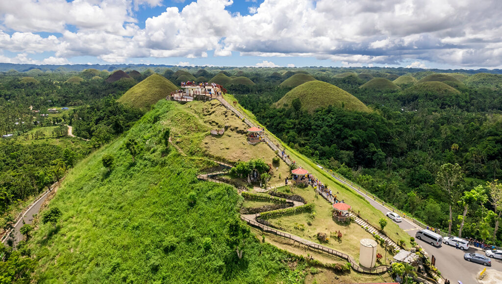 A high-angle view of a main viewing deck atop one of the Chocolate Hills Bohol, featuring a long concrete staircase with several covered rest areas leading up the grassy slope. Tourists are seen on the observation platform and stairs, overlooking a vast landscape of iconic dome-shaped hills and lush green tropical forests under a bright, cloudy sky.