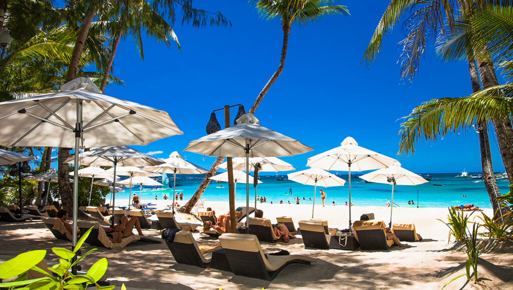 Vacationers relaxing on comfortable sun loungers under white beach umbrellas on a tropical shore, showcasing the premium beachfront amenities of family resorts in Philippines.