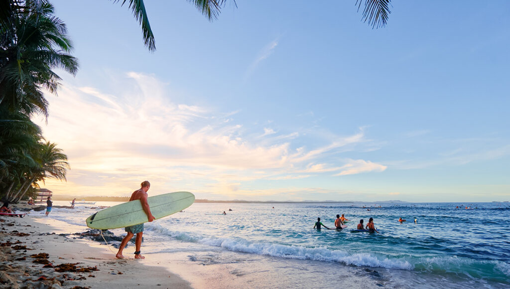 A surfer carrying a longboard walks toward the ocean at sunset, capturing the relaxed lifestyle at popular Siargao surf spots.