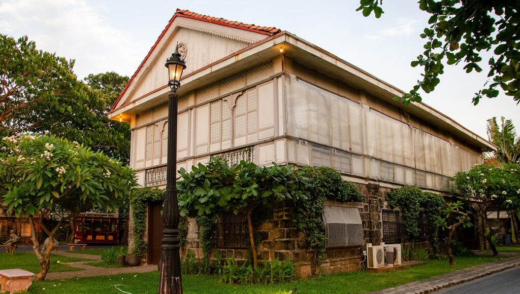 A beautiful two-story Bahay na Bato heritage house at Las Casas Filipinas de Acuzar, one of the premier historical places in Bataan. The structure features a dark stone ground floor, a white wooden upper level with capiz windows, and a red-tiled roof. A vintage black lamp post illuminates the foreground, set against a backdrop of lush green trees and a well-maintained lawn.