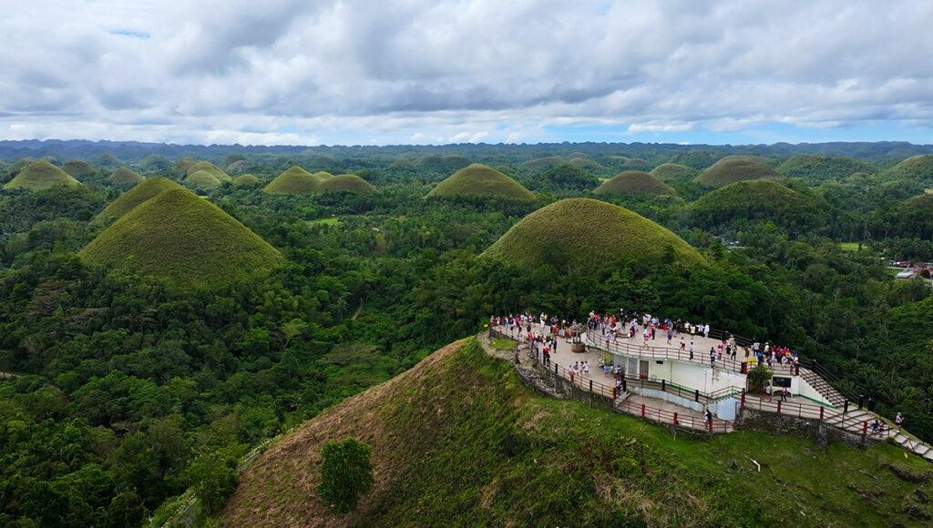 A panoramic view from the summit of one of the Chocolate Hills Bohol, featuring a large, white viewing platform filled with tourists. The platform overlooks a vast valley of symmetrical green hills and dense tropical forests under a dramatic, cloudy sky, capturing the immense scale of the geological formation.