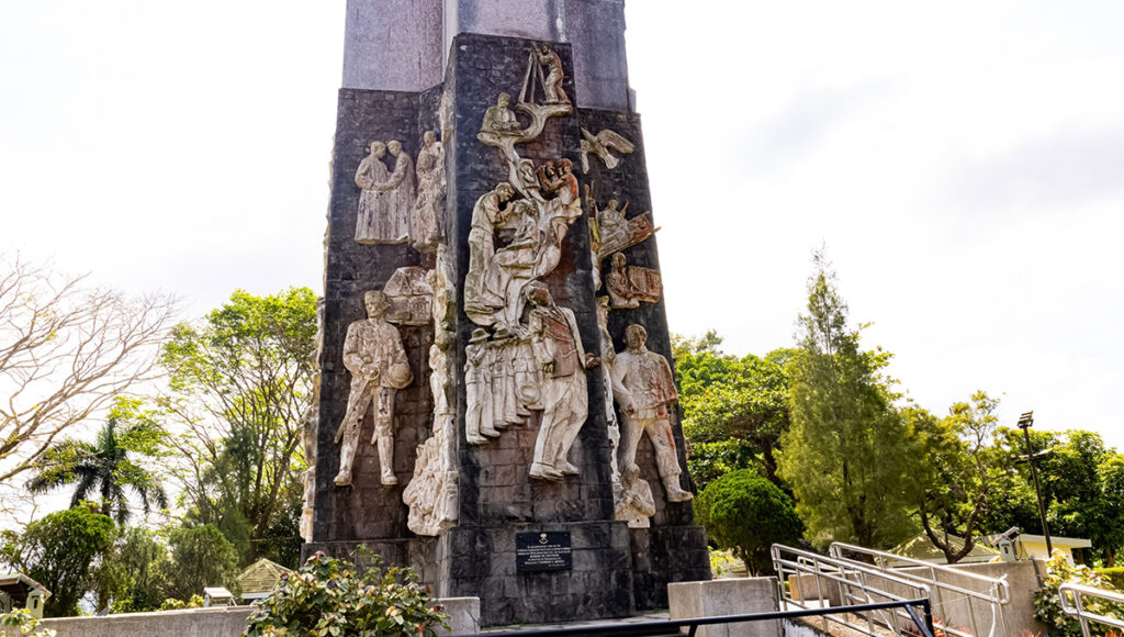 A detailed close-up of the intricate stone bas-relief carvings at the base of the Mount Samat Memorial Cross, a landmark among historical places in Bataan. The sculptures depict scenes of heroism and the struggles of soldiers and civilians during World War II. The dark stone monument is surrounded by green foliage and a clear sky, highlighting the craftsmanship of the National Shrine.