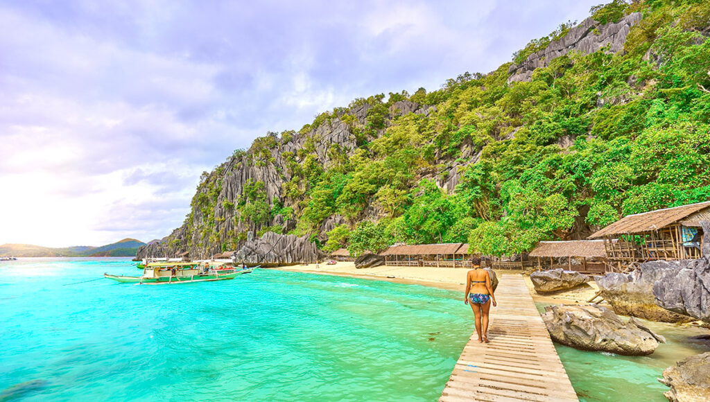 A wooden boardwalk leading to a white sand cove nestled against towering cliffs at one of the Palawan beaches.