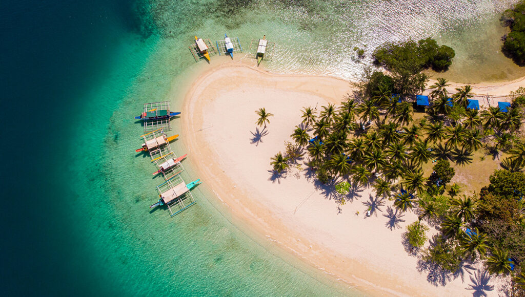 Aerial view of a white sandbar with palm trees and colorful boats, representing the remote beauty that influences how long to spend in the Philippines.