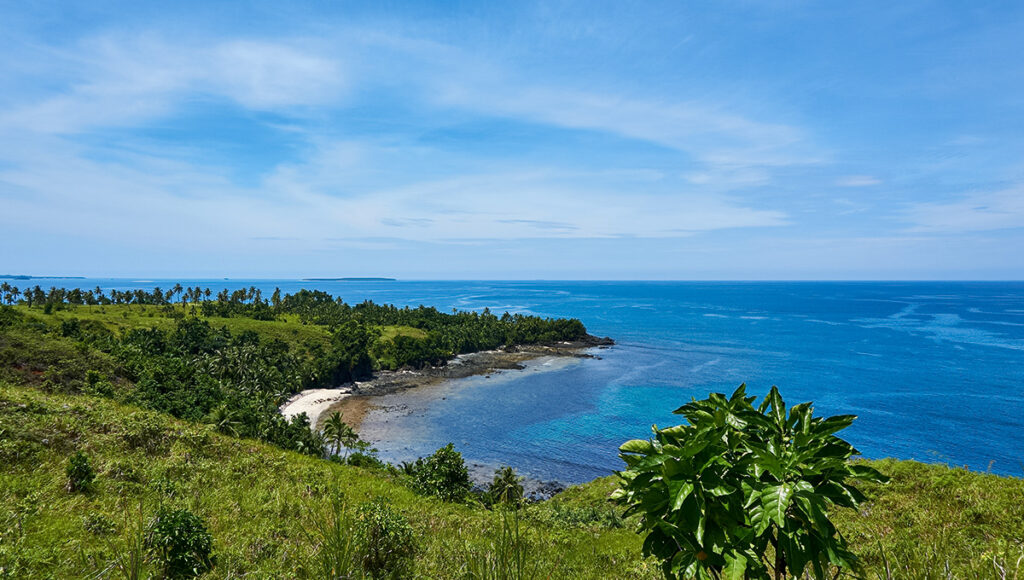 A wide scenic view of the lush, green cliffs and clear blue waters of the Bataan peninsula, home to many historical places in Bataan. The coastline features a small sandy cove tucked between rocky shores and tropical foliage, overlooking the vast sea under a bright, wispy sky—a landscape that served as the backdrop for the defense of the Philippines.