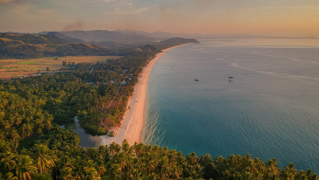 A high-angle sunset view of a long, sweeping coastline and lush palm forests bordering Palawan beaches.