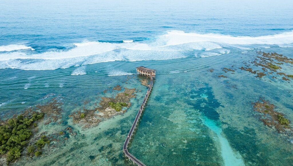 An aerial view of the iconic Cloud 9 boardwalk stretching over a vibrant coral reef toward the breaking waves, a central landmark for visitors at world-famous siargao surf spots.