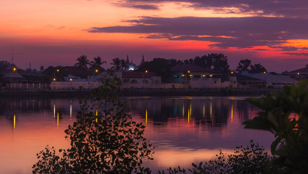 A vibrant sunset over the Iloilo River Esplanade, showing golden and purple hues reflected in the water with city lights beginning to glow, illustrating the relaxing things to do in Iloilo City after dark.
