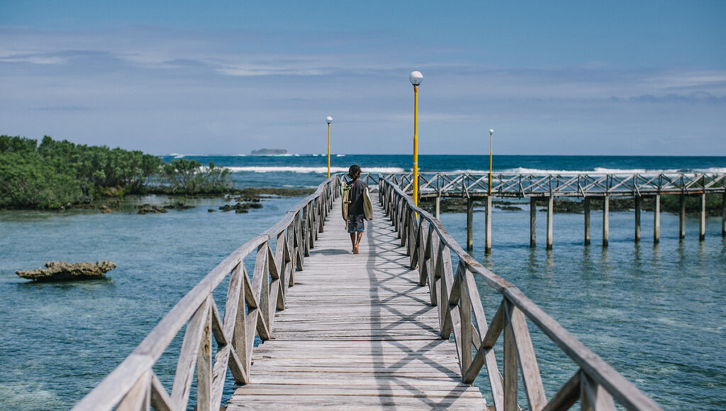 A lone surfer walking down the iconic Cloud 9 wooden boardwalk toward the ocean under a clear blue sky in Siargao.