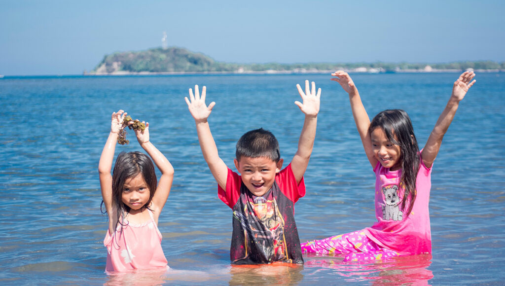 Local children playing in the turquoise shallows of a tropical beach, enjoying the bright sunshine and clear skies of the Philippines weather in March.