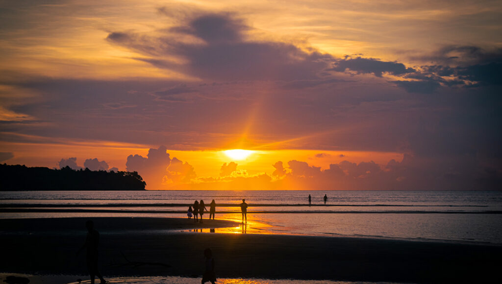A serene beach at sunset with several people silhouetted against a vibrant orange and yellow sky as the sun dips toward the horizon.