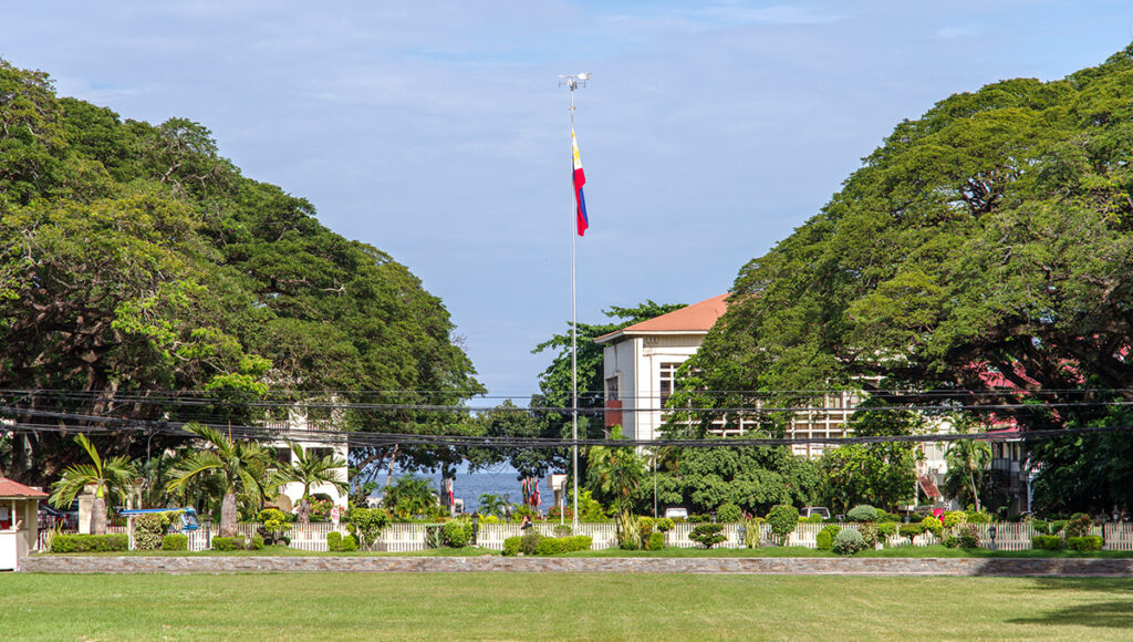 The Philippine flag flying high on a grassy field framed by giant acacia trees, showcasing cultural things to do in Dumaguete.