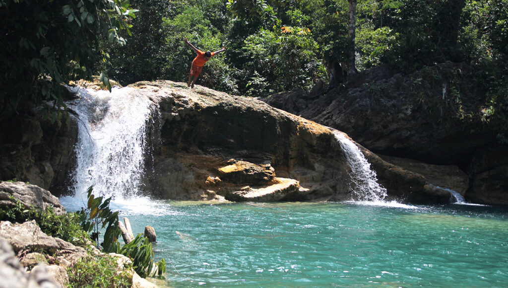 A young man jumping off a rock ledge into the turquoise pool of Bao-Bao Falls, one of the top adventure-filled places to visit in Surigao. The image captures two waterfalls cascading over smooth rocks into clear blue water surrounded by lush forest.