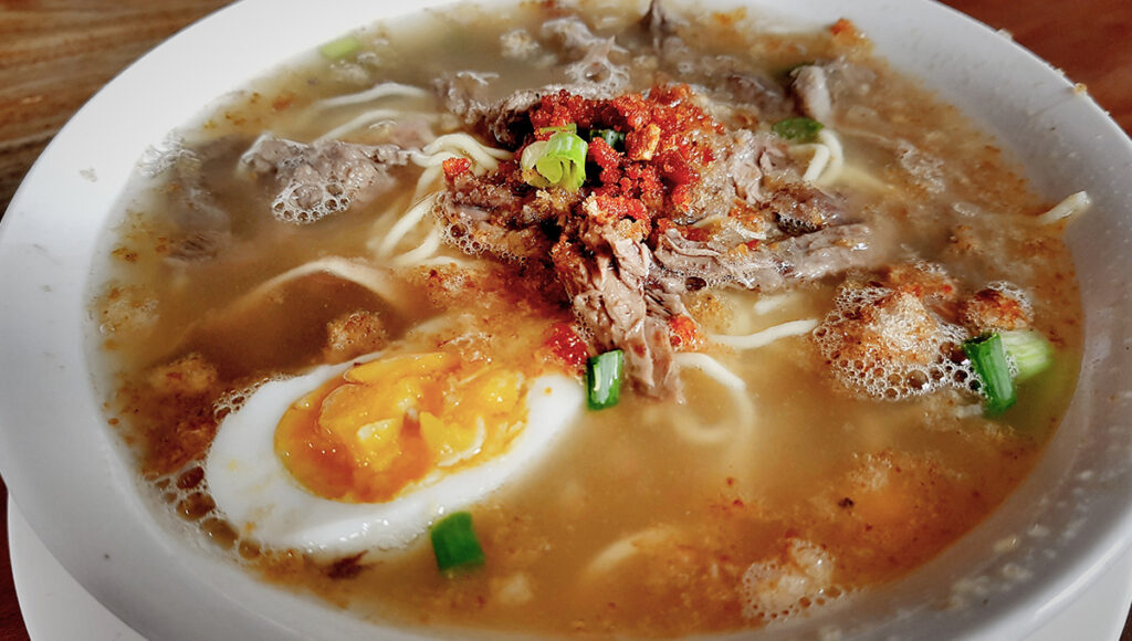 A steaming bowl of authentic La Paz Batchoy featuring egg noodles, sliced pork, crushed chicharon, and a soft-boiled egg in a rich broth—one of the most iconic culinary things to do in Iloilo City.