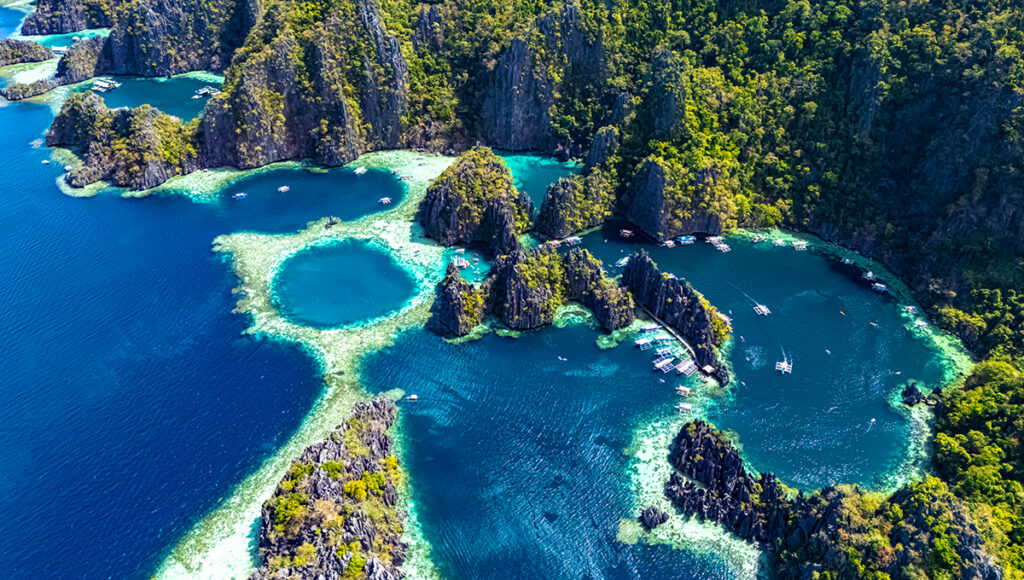 n aerial view of a tropical lagoon with turquoise water, limestone cliffs covered in greenery, and several small boats anchored near coral reefs.