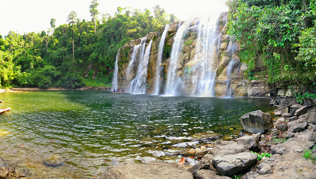 The majestic Tinuy-an Falls in Bislig, a premier destination among places to visit in Surigao. This wide, multi-tiered waterfall cascades into a large natural pool surrounded by a dense green rainforest, with a bamboo raft visible near the base.