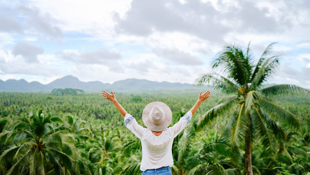A woman with her arms raised in a wide-brimmed hat overlooking a vast, endless forest of coconut palm trees in Siargao.
