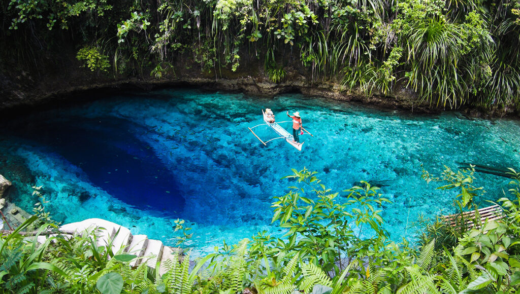 The tranquil, turquoise waters of Bogac Cold Spring in Barobo, one of the most refreshing places to visit in Surigao. Lush green trees and coconut palms surround the crystal-clear natural swimming hole under a bright blue sky.
