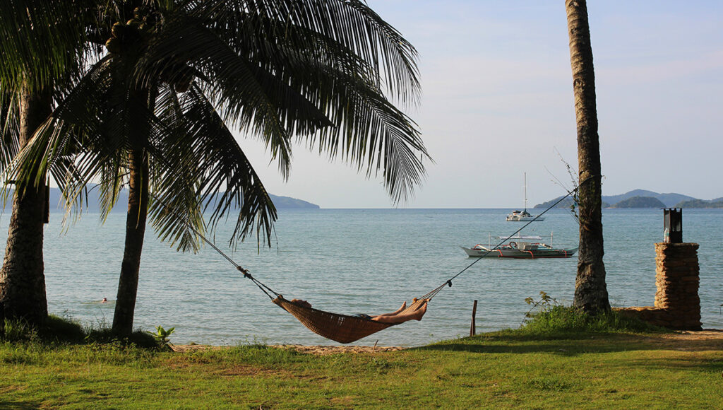 A traveler relaxing in a hammock between palm trees, enjoying a coastal breeze to beat the intense heat of the Philippines weather May.