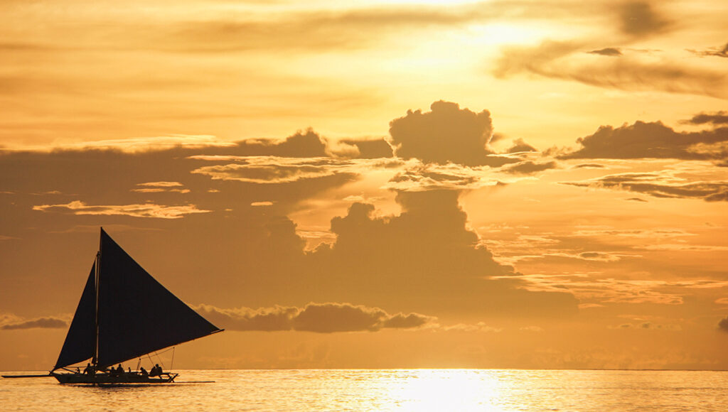 A traditional sailboat with a dark triangular sail gliding across a calm sea during a golden sunset with heavy, dramatic clouds.