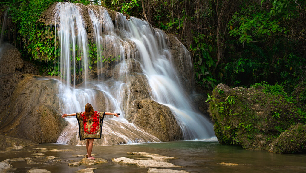 A woman in a red patterned dress standing with her arms outstretched in front of a wide, multi-tiered waterfall surrounded by lush tropical forest.