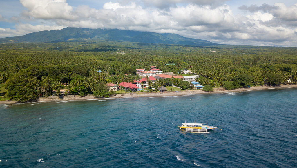 Aerial view of a beachfront resort with red roofs and a yellow boat anchored in the blue sea, highlighting leisure things to do in Dumaguete.