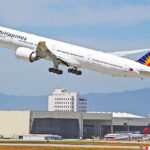 A Philippine Airlines Boeing 777-300ER, with its distinctive red, blue, and yellow tail fin, is captured in mid-takeoff against a clear sky with distant mountains. This image captures the excitement of air travel, which brings up the key question for many travellers: When will Philippine Airlines resume flights to London? The aircraft is angled upwards, with its landing gear still extended. Below, airport buildings and a runway are visible, along with other aircraft tails in the background.