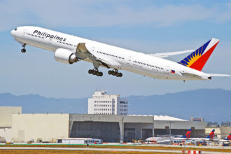 A Philippine Airlines Boeing 777-300ER, with its distinctive red, blue, and yellow tail fin, is captured in mid-takeoff against a clear sky with distant mountains. This image captures the excitement of air travel, which brings up the key question for many travellers: When will Philippine Airlines resume flights to London? The aircraft is angled upwards, with its landing gear still extended. Below, airport buildings and a runway are visible, along with other aircraft tails in the background.