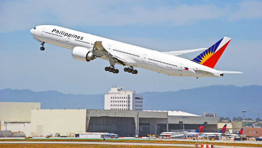 A Philippine Airlines Boeing 777-300ER, with its distinctive red, blue, and yellow tail fin, is captured in mid-takeoff against a clear sky with distant mountains. This image captures the excitement of air travel, which brings up the key question for many travellers: When will Philippine Airlines resume flights to London? The aircraft is angled upwards, with its landing gear still extended. Below, airport buildings and a runway are visible, along with other aircraft tails in the background.