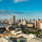 Panoramic view of the modern Manila skyline and waterfront, showing skyscrapers, a marina, and an amusement park with a Ferris wheel in the foreground, suggesting a vibrant and developed city. This image helps answer the question, "Is Manila safe?" by illustrating a bustling, modern urban center.