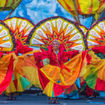 An image of three Filipino women in vibrant red, yellow, and orange traditional festival attire, performing a street dance with large, colorful, sun-like fan backdrops. The photo captures the energy and bright colors of Filipino culture.