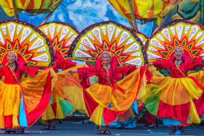 An image of three Filipino women in vibrant red, yellow, and orange traditional festival attire, performing a street dance with large, colorful, sun-like fan backdrops. The photo captures the energy and bright colors of Filipino culture.