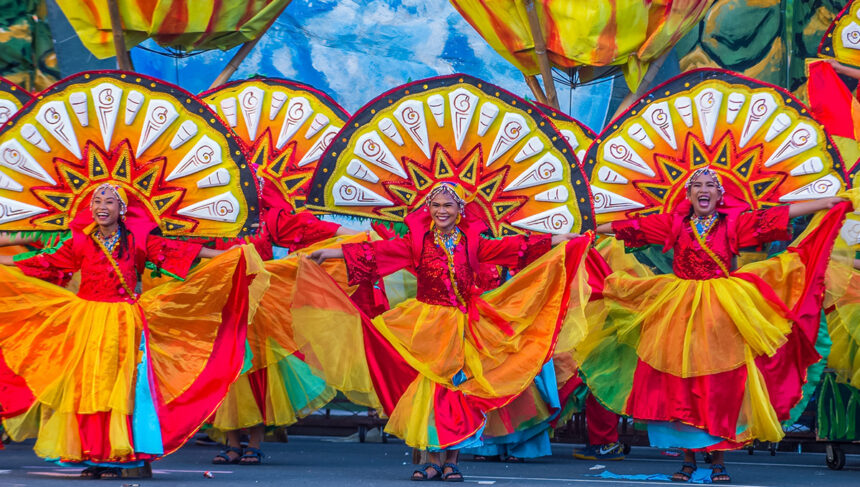 An image of three Filipino women in vibrant red, yellow, and orange traditional festival attire, performing a street dance with large, colorful, sun-like fan backdrops. The photo captures the energy and bright colors of Filipino culture.