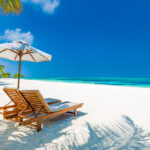 A beautiful tropical beach scene featuring two wooden lounge chairs and a white umbrella on pristine white sand, with palm leaves casting shadows, bright turquoise ocean water, and a clear blue sky, evoking the atmosphere of all inclusive resorts Philippines.