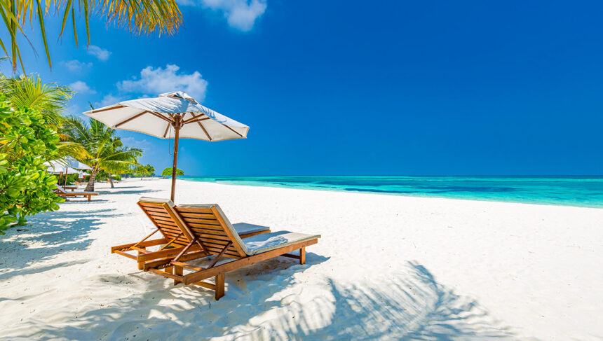 A beautiful tropical beach scene featuring two wooden lounge chairs and a white umbrella on pristine white sand, with palm leaves casting shadows, bright turquoise ocean water, and a clear blue sky, evoking the atmosphere of all inclusive resorts Philippines.