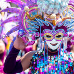 use the keyword , masskara festival in bacolod city and make an alt text for this image. A person adorned in a vibrant, elaborate costume and mask, characteristic of the MassKara Festival in Bacolod City, Philippines, is shown from the chest up. The mask features a smiling face with intricate designs in gold, purple, and blue, framed by large, feathery purple and pink adornments. The costume includes a sequined top in shades of blue, purple, and gold, and the person's arms are covered in purple sleeves, holding the mask in place. The background is blurred, showing other costumed participants, suggesting a festive parade or celebration under bright daylight.
