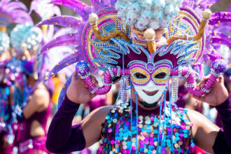 A person adorned in a vibrant, elaborate costume and mask, characteristic of the MassKara Festival in Bacolod City, Philippines, is shown from the chest up. The mask features a smiling face with intricate designs in gold, purple, and blue, framed by large, feathery purple and pink adornments. The costume includes a sequined top in shades of blue, purple, and gold, and the person's arms are covered in purple sleeves, holding the mask in place. The background is blurred, showing other costumed participants, suggesting a festive parade or celebration under bright daylight.